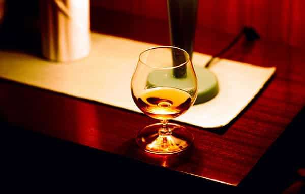 Auf einer Ablage aus Mahagoni in einem Hotel in Frankreich steht ein gefuelltes Cognacglas. Die Szene wird durch eine kleine Lampe beleuchtet und taucht sie in ein warmes Licht, so das der Cognac guelden illuminiert ist. Die kleine Tischlampe steht auf einer laenglichen weissen Decke, ihr Fuss ist gruen, der Stab dunkelbraun. Den Lampenschirm sieht man nicht. Eine weisse Vase steht im Anschnitt links von der Lampe