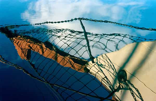 Alles nur Spiegelungen in einem dunkelblauen Wasser. Da ist der beige Rumpf mit seinem Pulpit, der mit braunem Segeltuch ummantelt ist, die weissen Quellwolken. Ein Netz und eine metallische Gliederkette sind durch die Wellenbewegung des Meeres verschwommen, verlieren sich teilweise im Himmel. Der Aufnahmeort ist Menorca