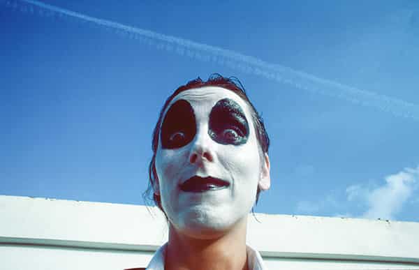 Ein junges Frauengesicht ist clownesk weiss geschminkt. Ihre Augen sind nach oben steigend oval schwarz umrandet, ihre Lippen sind ebenfalls schwarz. Der Hintergrund besteht zu vier fuenfteln aus blauem Himmel mit wenig weissem Wolkenanteil und ein Kondensstreifen geht ueber die gesamte Bildbreite. Aufnahmeort ist Menorca
