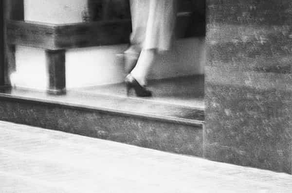 Das Bild ist unscharf, die Kamera geschwenkt, die Verschlusszeit lang. Eine Lady hat die Schwelle aus Marmor bereits ueberwunden und ihr linkes Bein macht den Schritt nach vorne in einen Eingang. Sie traegt einen bis ueber die Knöchel reichenden Rock und schwarze Poemps. Die Fassade ist aus Marmor und im linken Bereich ist eine Holzkonstruktion zu sehen. Der Buergersteig  verlaeuft diagonal, ist hell und natuerlich auch unscharf. Es ist eine Schwarz - Weiss - Aufnahme, entstanden in Frankfurt.