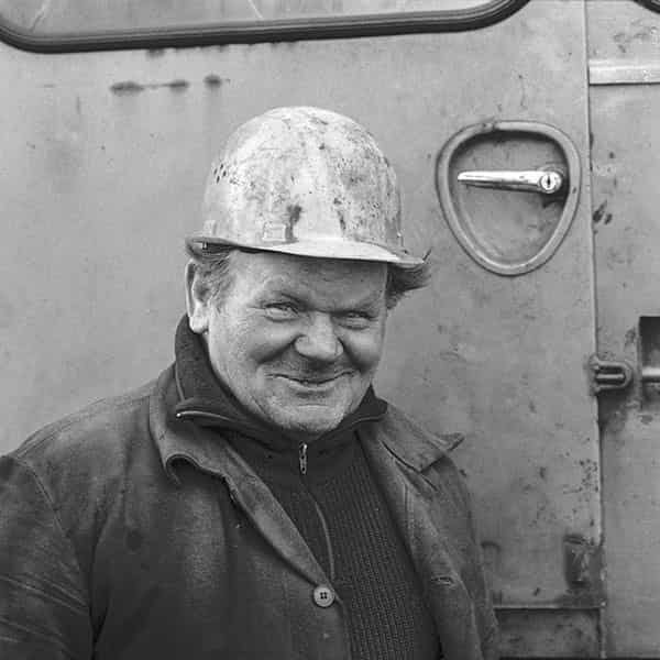 Ein Sinnbild des Arbeiters; zusammen gekniffene Augen, die lachen - der Mund ein breites Grinsen. Bartstoppeln und ein Helm auf dem Kopf, unter dem sich Haarbueschel heraus nach oben winden. Die Knollennase sticht hervor. Er ist dem kalten Februar in Hamburg entsprechend mit einer Strickjacke, worueber  er eine grobe Lederjacke traegt, bekleidet. Der Hintergrund ist die Tuer zu einem Bagger und der Griff zum Eintritt befindet sich in Helmhoehe.