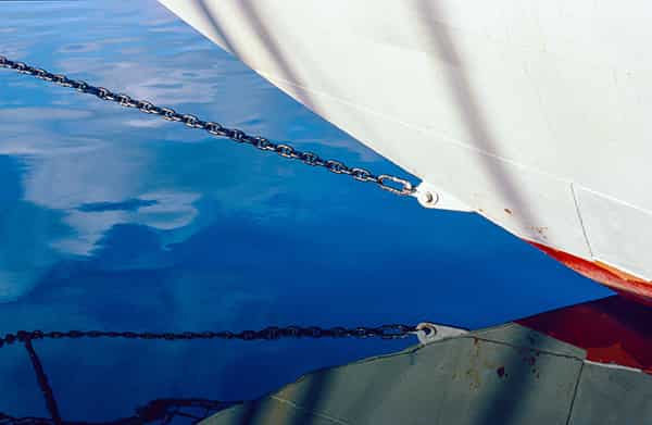 Erneut der Bug eines Schiffes mit seinen Spiegelungen im blauen Meerwasser. Die Farbe des Wassers changiert von tiefem Blau bis hin zu Weiss mit einem leichten Rotstich. Eine eiserne Gliederkette sieht man sowohl oberhalb der Wasseroberflaeche, sowie als Spiegelung, welche durch eine sanfte Meeresbewegung teilweise unscharf ist. Der reale Bug ist weiss und hat, bevor er in das Wasser eintaucht, einen roten Anstrich, den man nur als kleinen Flecken im Anschnitt sieht. Die Spiegelung intensiviert die Farben. Der Aufnahmeort ist Menorca