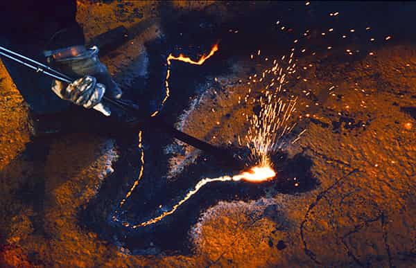 Im zweiten Bild der Serie sieht man bereits die Kontur vom Wolfkopf als eine Linie aus Feuer. Eine Hand, mit einem Handschuh geschuetzt, ragt in das Bild und fuehrt eine Schweisslanze.  Die Farben sind orange, dunkelblau, Feuer und Silber. Aufgenommen wurde das Foto in einem Abwrackbetrieb im Hafen von Hamburg.