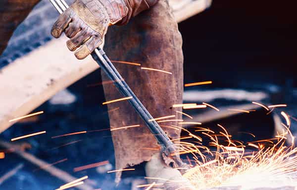 Die Funken spruehen in den Farben von weissgelb ueber gelb, gelborange bis zu orange. Man sieht ein Bein, mit einem Lederschutz vor den Funken. Eine starke Hand umgreift die Schweisslanze. Sie wird durch einen Handschuh vor den heissen Funken geschuetzt. Aufgenommen wurde das Foto in einem Abwrackbetrieb im Hafen von Hamburg.